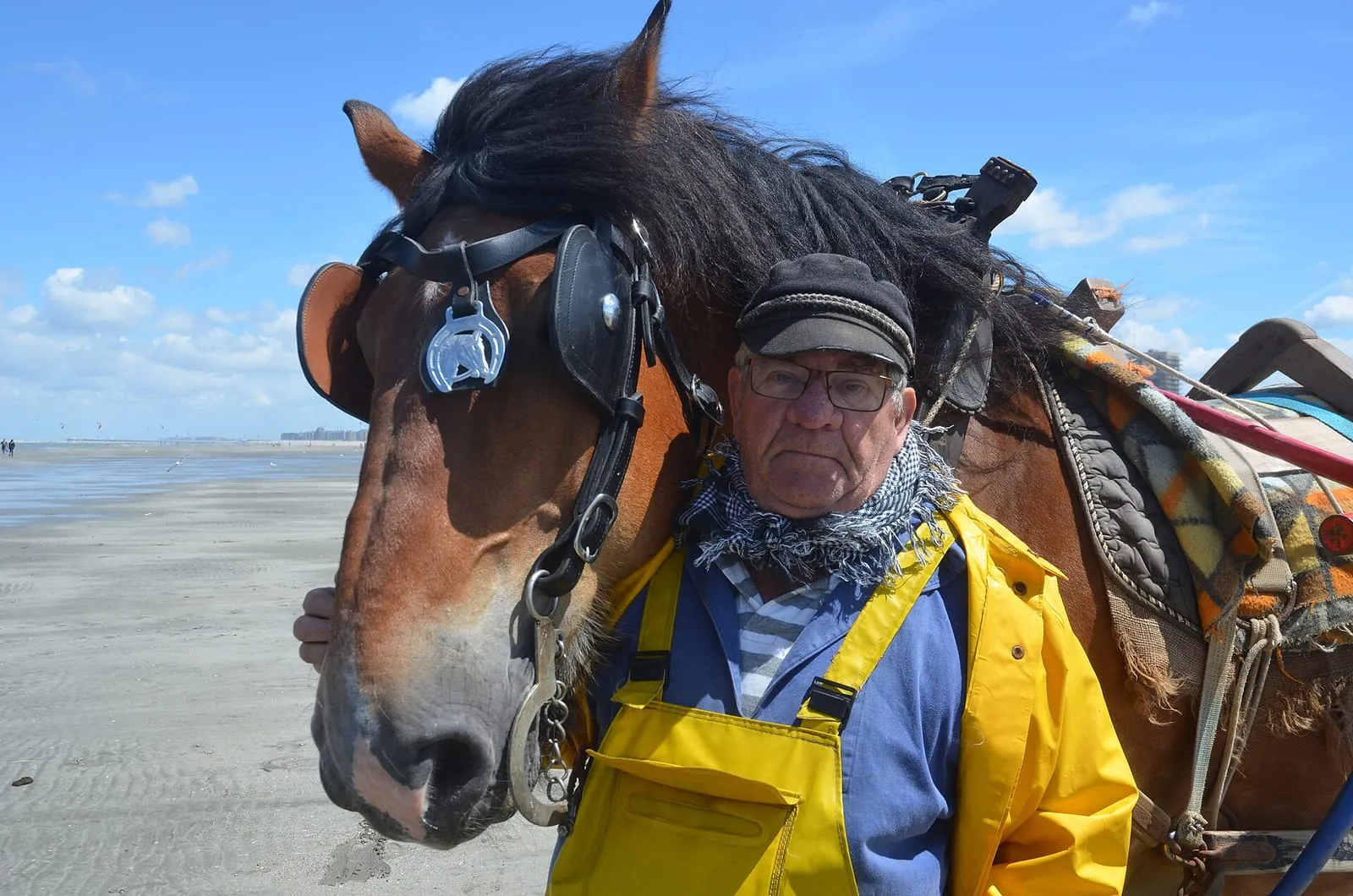 Een garnaalvisser met pet en gele overall staat tegen zijn paard aan op het strand