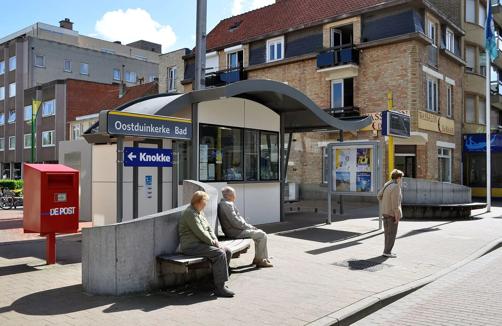 De kusttramhalte Oostduinkerke Bad met een overkapping, een bank, een rode brievenbus en een huis erachter