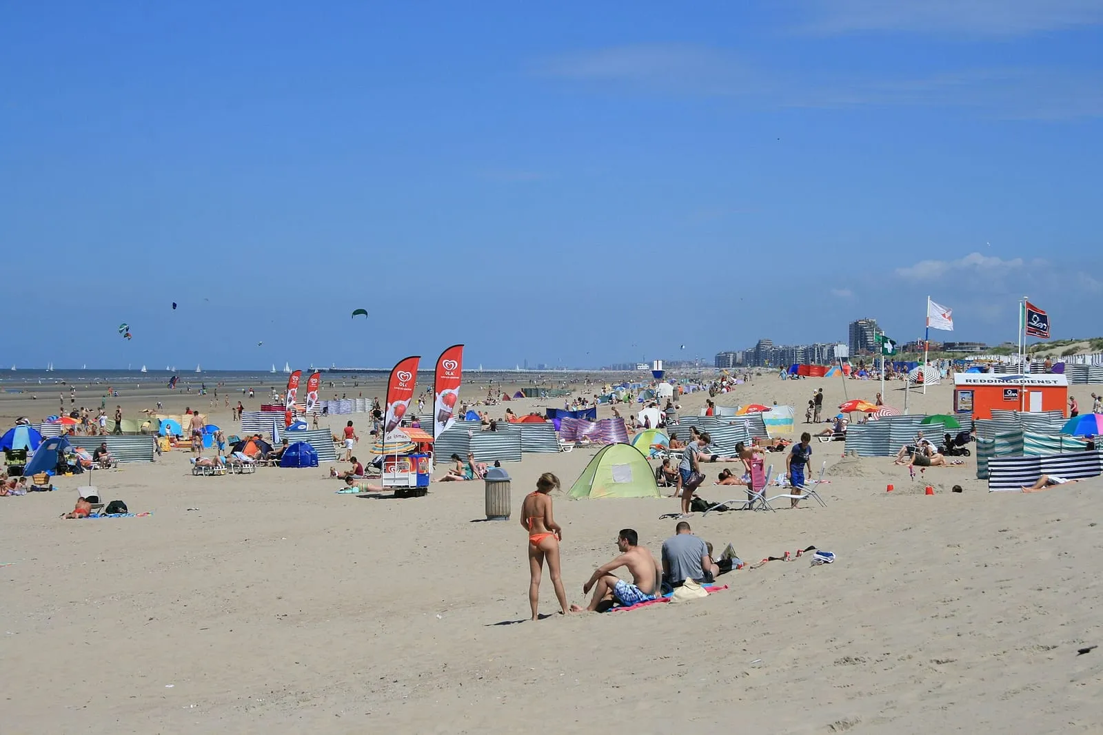 Breed zandstrand met badgasten en kleurrijke windschermen onder een blauwe lucht, de zee in de verte