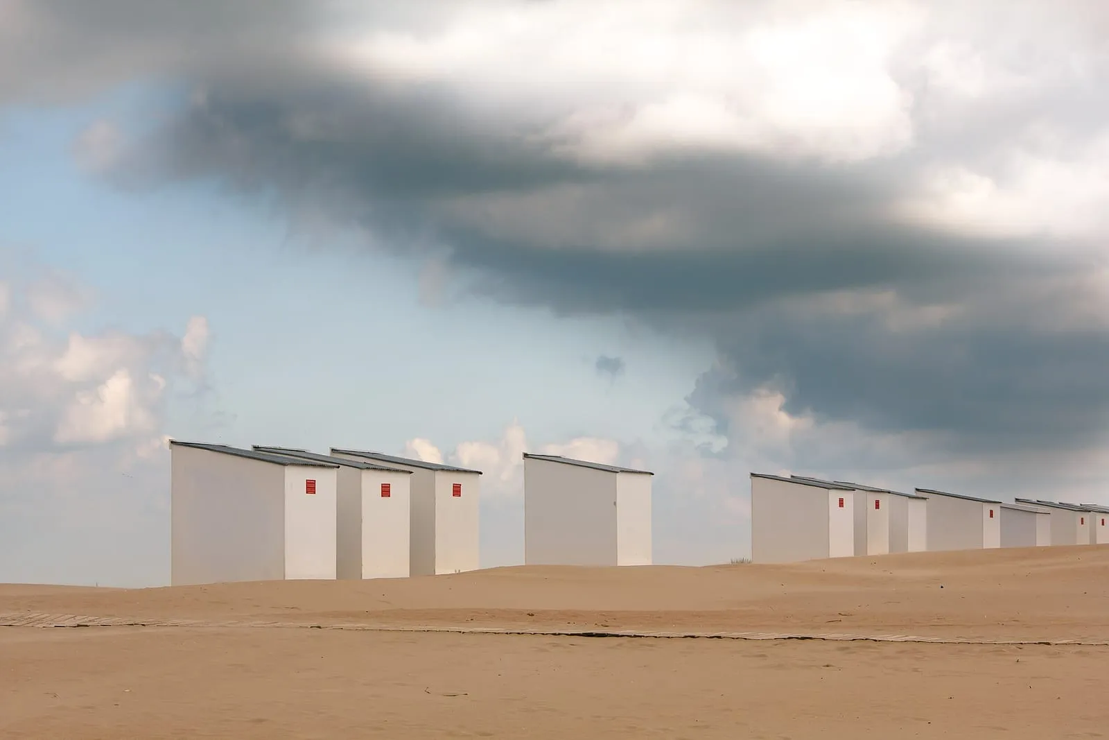 Een rij witte strandcabines op het zand onder een zware, grijze wolkenlucht