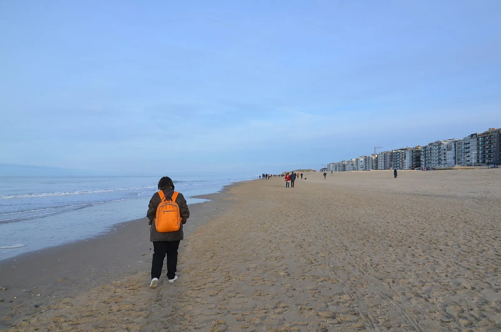Een wandelaar met een oranje rugzak loopt over een breed strand onder een blauwe lucht, met flatgebouwen in de verte