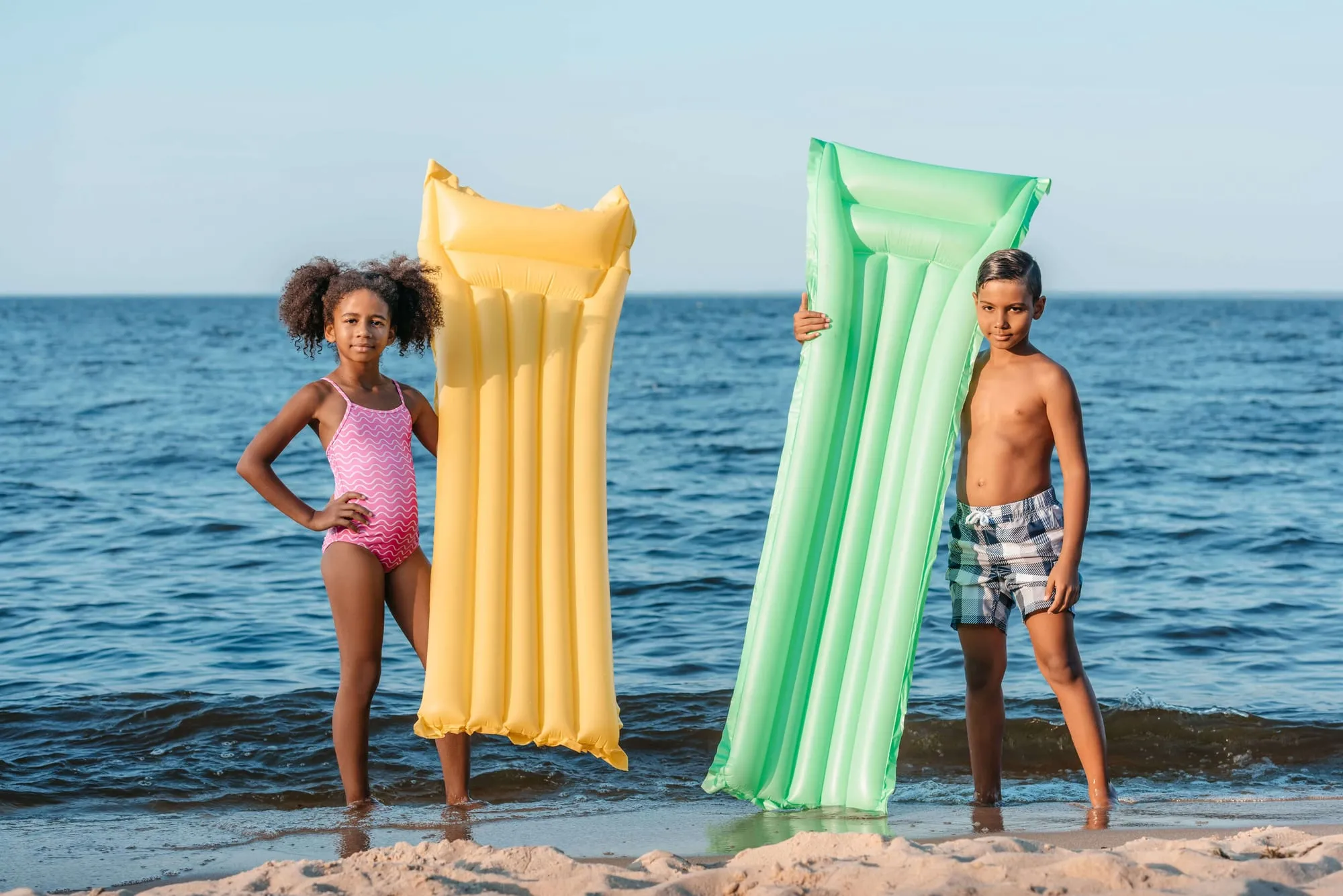 Twee kinderen staan op het strand met een geel en een groen luchtbed, de zee achter hen
