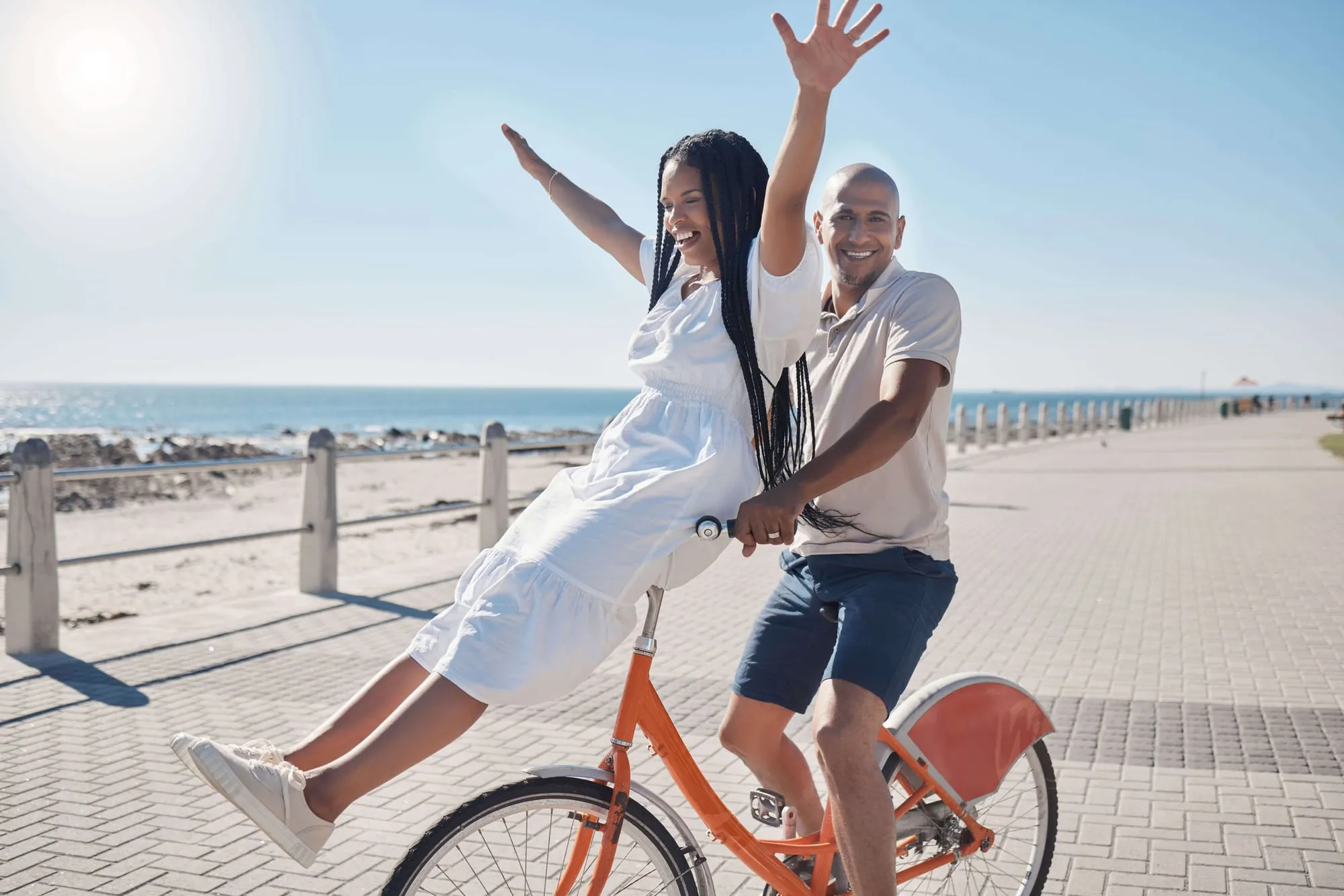 Een lachend stel op één fiets op een brede promenade aan zee, de vrouw steekt haar armen in de lucht