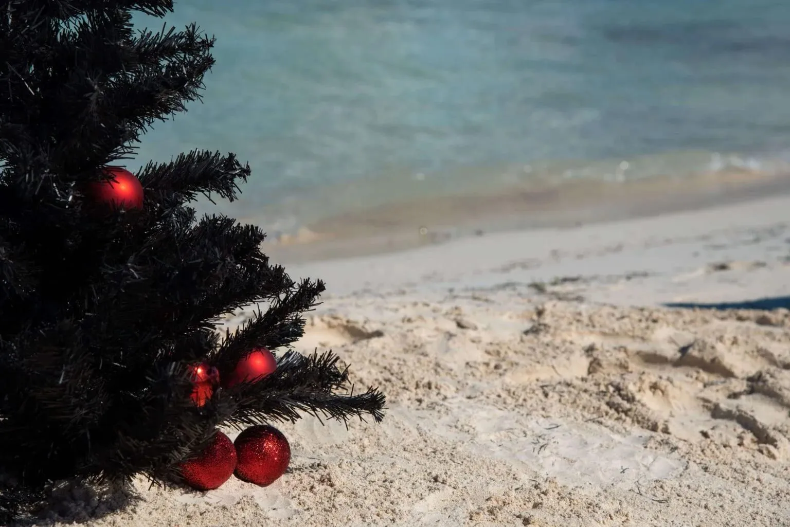 Een kerstboom met rode kerstballen op een zandstrand, met de zee op de achtergrond