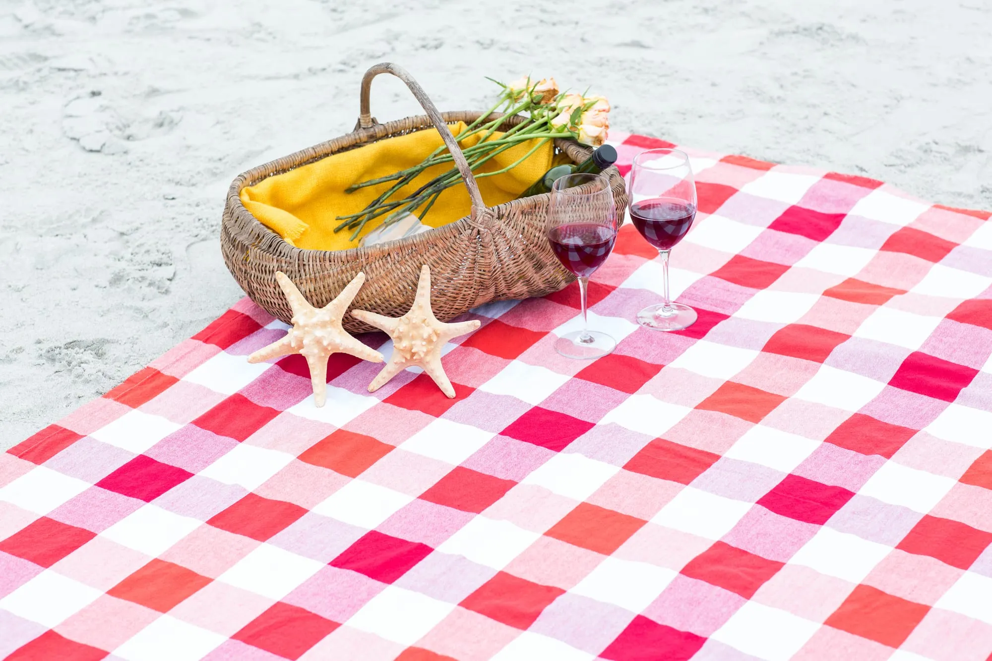 Een rieten picknickmand en twee glazen rode wijn met zeesterren op een rood-wit geruit kleed op het strand