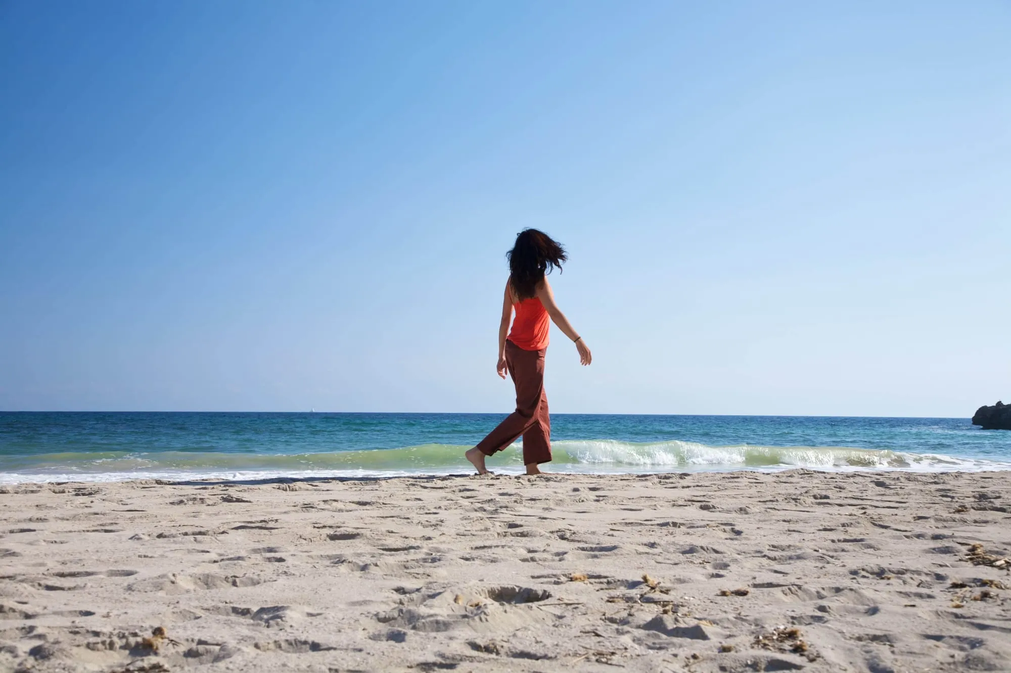 Een vrouw wandelt over een breed, leeg zandstrand onder een blauwe lucht